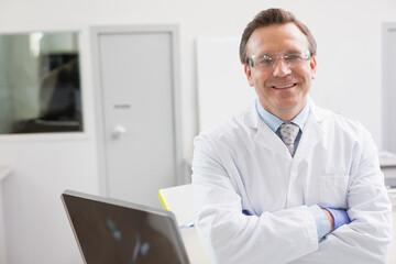 Middle-aged man in lab coat crossing arms at workbench with laptop and safety glasses, copy space