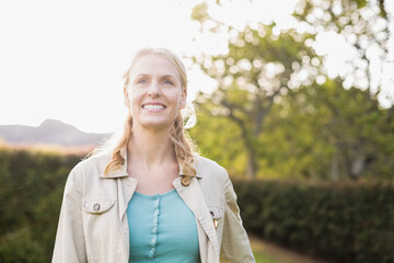 Woman in her 30s smiling and looking upward while standing in sunlit garden wearing beige jacket