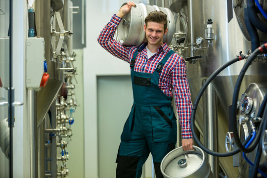 Male brewery worker wearing overalls handling beer kegs between steel tanks with valves and hoses