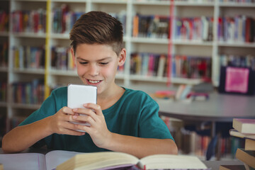 School-age boy sitting at library table holding smartphone reading open book with stacked books