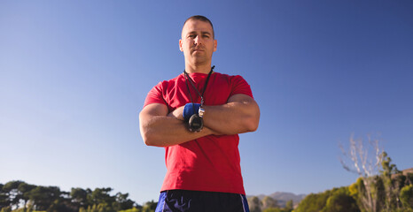 Male fitness coach standing with arms crossed in sportswear on field with stopwatch, wrist wrap