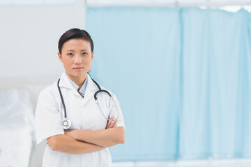 Doctor wearing white coat and stethoscope standing with arms crossed by exam bed, privacy curtain