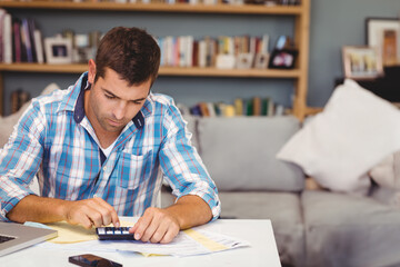 Man sitting at table in living room using calculator with laptop phone and documents, copy space