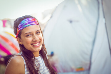 Woman smiling at camera standing among canvas festival tents in tie-dye headband, striped tank top