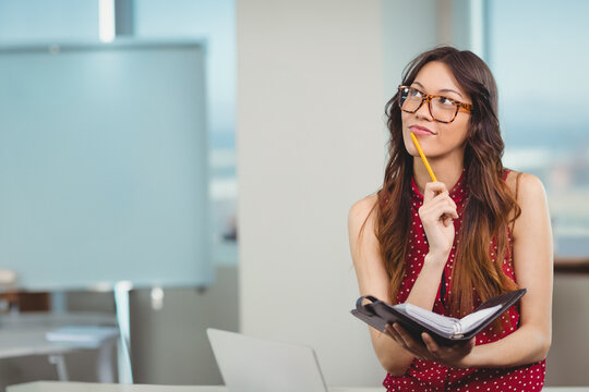 Businesswoman sitting in office holding planner and pencil beside laptop and whiteboard, copy space - Powered by Adobe