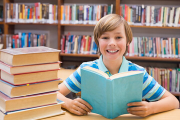 Preteen student reading light blue book at wooden library table, with stacked volumes behind