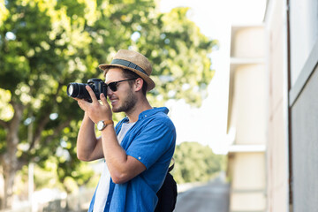 Photographer carrying backpack, aiming DSLR camera on sidewalk by light-colored building wall