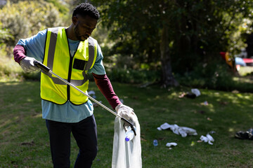 African American man picking up litter in park with metal picker and plastic-lined bag, copy space