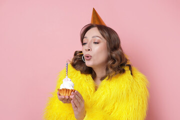 Birthday celebration. Woman blowing candle on cupcake against pink background
