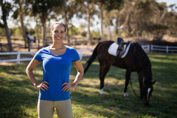 Smiling woman in riding gear standing in grassy paddock near white fence with saddled horse grazing