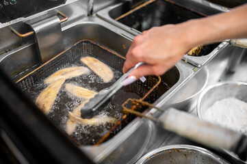 A close-up of a chef using tongs to fry fish fillets in hot oil. The bubbling oil surrounds the fillets, capturing the texture and movement. The scene is set in a professional kitchen