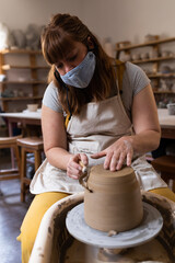 Clay vessel spinning on pottery wheel in ceramics studio, with wooden shelves holding bisque pieces