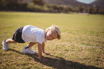 Boy crawling in plank pose on grass sports field wearing sneakers under sunshine, copy space