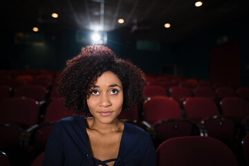 African American woman sitting facing screen in dim theater with red seats and projector light