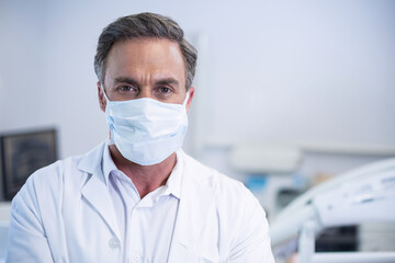 Male doctor standing in examination room wearing lab coat and surgical face mask