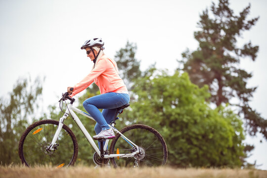 Fototapeta Woman riding white mountain bicycle up grassy hill at forest edge wearing helmet and sunglasses