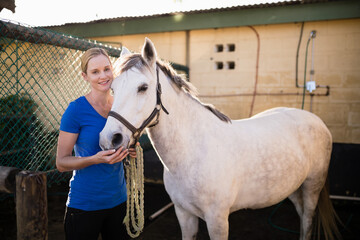 Woman holding braided lead rope touching white horse muzzle in stable yard by chain link fence