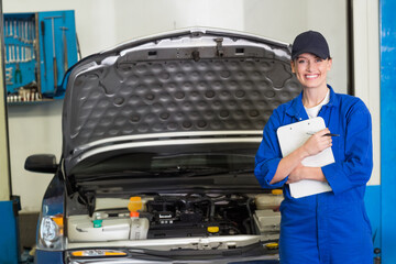 Female mechanic in blue coveralls checking open car hood in workshop holding clipboard, copy space