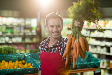 Male produce clerk smiling and presenting fresh carrots beside mesh orange bins at supermarket