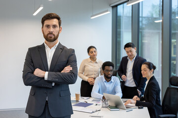 A confident businessman stands with arms crossed in the foreground, while his team collaborates around a table in a modern office setting.