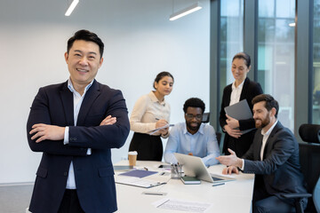 A confident Asian businessman smiles with arms crossed, leading a diverse team in a modern office environment.