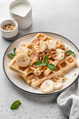 Belgian waffles with banana, nuts and honey on plate served with mint leaves on white table background. Sweet breakfast concept