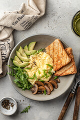 Scrambled eggs, toasts, avocado and mushrooms on rustic plate for homemade breakfast, top view