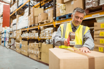 Senior male warehouse worker in safety vest taping box with tape dispenser in aisle, copy space