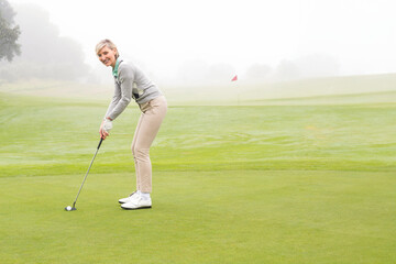 Fototapeta premium Middle-aged woman in golf gear gripping putter lining up golf ball on putting green with flagstick