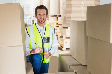 Male warehouse supervisor kneeling among pallets and boxes in storage facility writing on clipboard