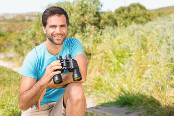 Man kneeling on dirt path in grassy field holding binoculars and smiling toward camera, copy space