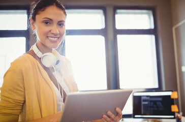 Hispanic woman holding tablet with headphones near desk monitor and orange notes at office windows