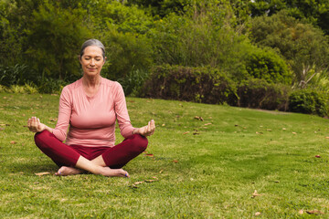 Senior woman meditating on grassy lawn in garden wearing pink athletic top and burgundy pants