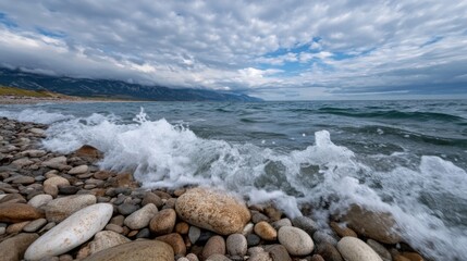 Waves roll onto a rocky shoreline, creating splashes among smooth stones. Dark clouds linger overhead, hinting at a dramatic sky while mountains are visible in the distance.