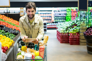 Mid adult male shopper pushing cart through supermarket produce aisle holding tablet and baguette