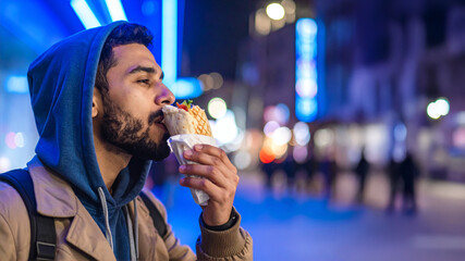 A person in a blue hoodie and beige jacket holds a wrapped sandwich while walking through a lively city at night. Bright neon lights and blurred figures in the background evoke themes of street food c