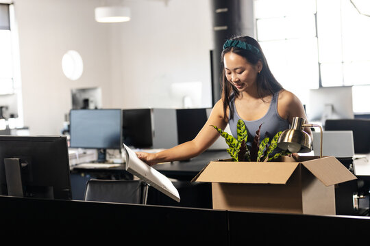 Chinese Asian woman unpacking box on desk in open-plan office placing lamp and plant, copy space