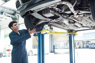 Male mechanic inspecting front wheel assembly under lifted car chassis at repair garage, copy space