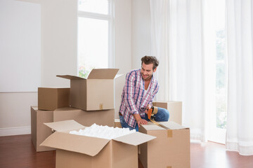 Man kneeling in room among foam peanuts and blank board, taping cardboard boxes with tape dispenser
