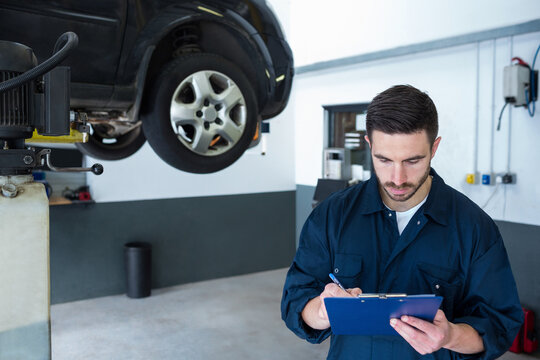 Male mechanic writing on blue clipboard beside car on hydraulic lift in repair garage