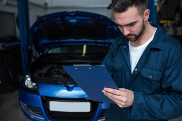 Adult male mechanic in blue coverall reading clipboard in repair garage with blue car, copy space
