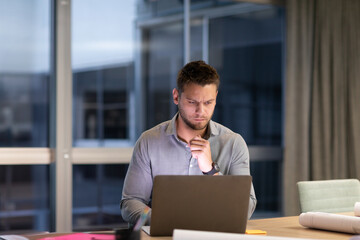 Mid adult male working on laptop at desk with architectural plans and folders in modern office