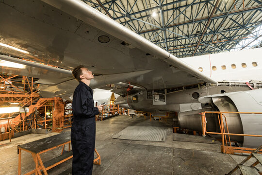 Male mechanic in uniform holding tablet, examining jet wing under scaffolding in hangar, copy space - Powered by Adobe