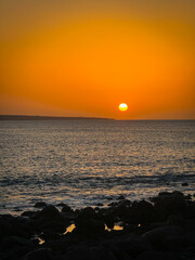 Brilliant orange sun dips towards horizon, casting a warm glow across the sky and reflecting on gently rippling ocean waves. Dark volcanic rocks create a silhouette against serene and fiery sunset.