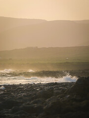 Waves crash against dark, rocky volcanic shore and disappear into a misty, ethereal haze that envelops distant hills. Muted tones and soft lighting create an atmospheric, mysterious coastal landscape.