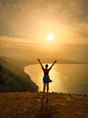 SILHOUETTE, LENS FLARE: Woman stands on clifftop with her arms outstretched, enjoying breathtaking view of Famara beach at sunrise. Morning sun creates a dramatic coastal view that enchants young lady