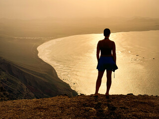 SILHOUETTE Female traveller on a clifftop gazing out at Famara beach and Atlantic Ocean shimmering under a soft, hazy light of golden sunset. Dramatic coastal landscape at volcanic island of Lanzarote