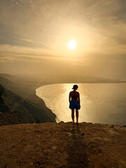SILHOUETTE, LENS FLARE: A lone female figure stands at a vantage point, observing a vast coastal bay framed by distant mountains. Stunning view from a clifftop of Famara beach in soft morning light.
