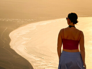 SILHOUETTE, CLOSE UP: Young woman gazes out at tranquil coastal bay, illuminated by a soft, ethereal light. Stunning clifftop view of Famara beach in golden light at volcanic island of Lanzarote.