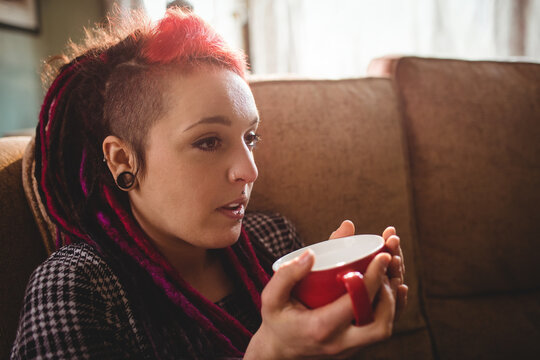 Woman in houndstooth sweater sitting on brown sofa at home holding red ceramic mug, copy space